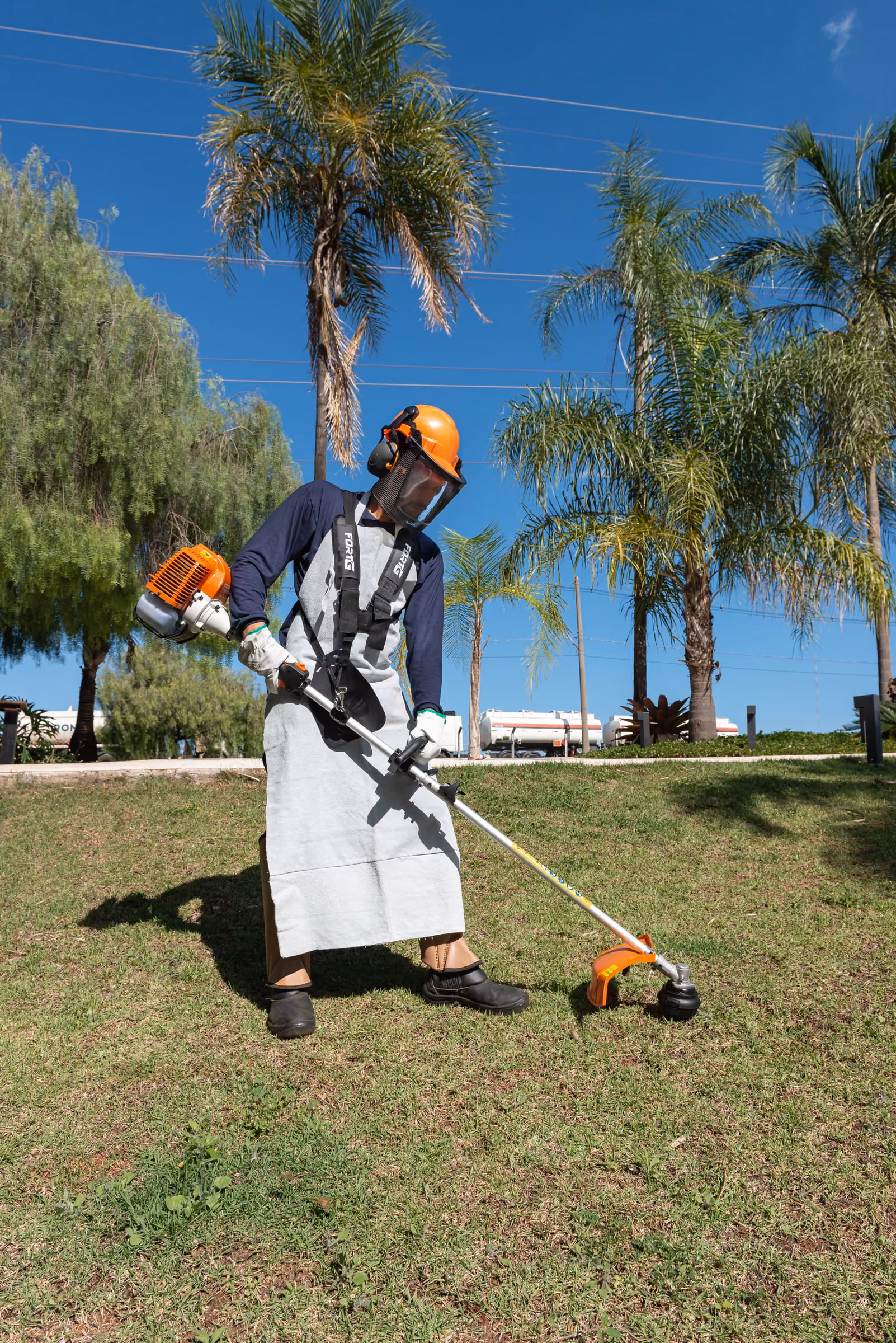 Jardineiro da Prátika com capacete, protetor facial, avental e luvas opera a roçadeira no gramado